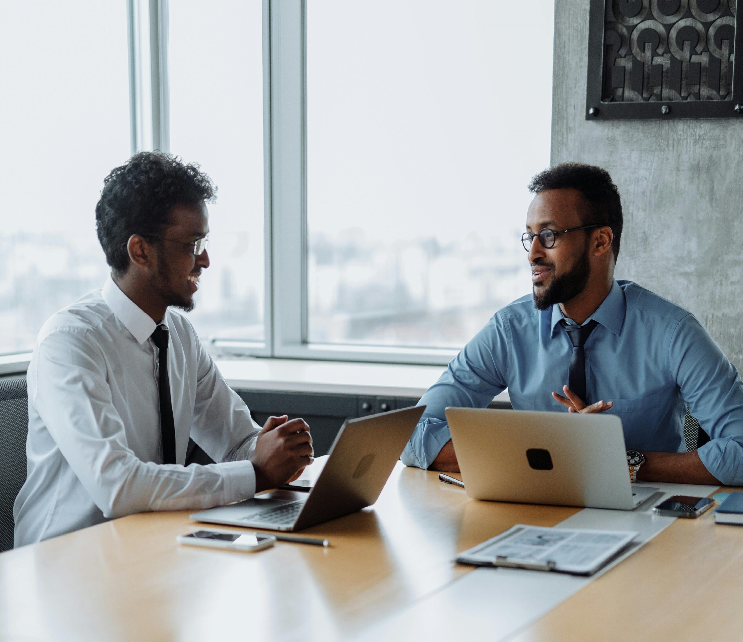 Two men in business attire discussing over laptops in a modern office setting.